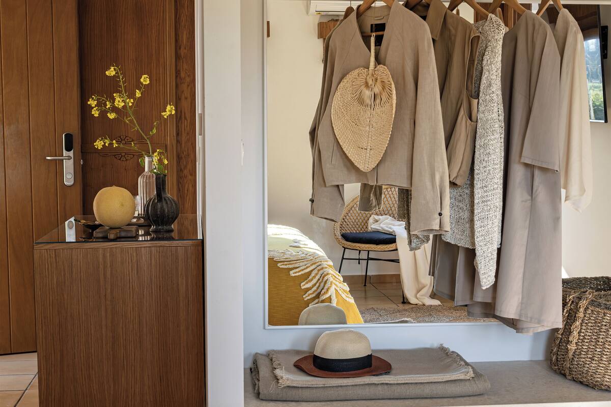 A room with a wooden wardrobe, a mirror reflecting clothes and a chair, a small table with a vase and a hat, and a bed with a yellow pillow.