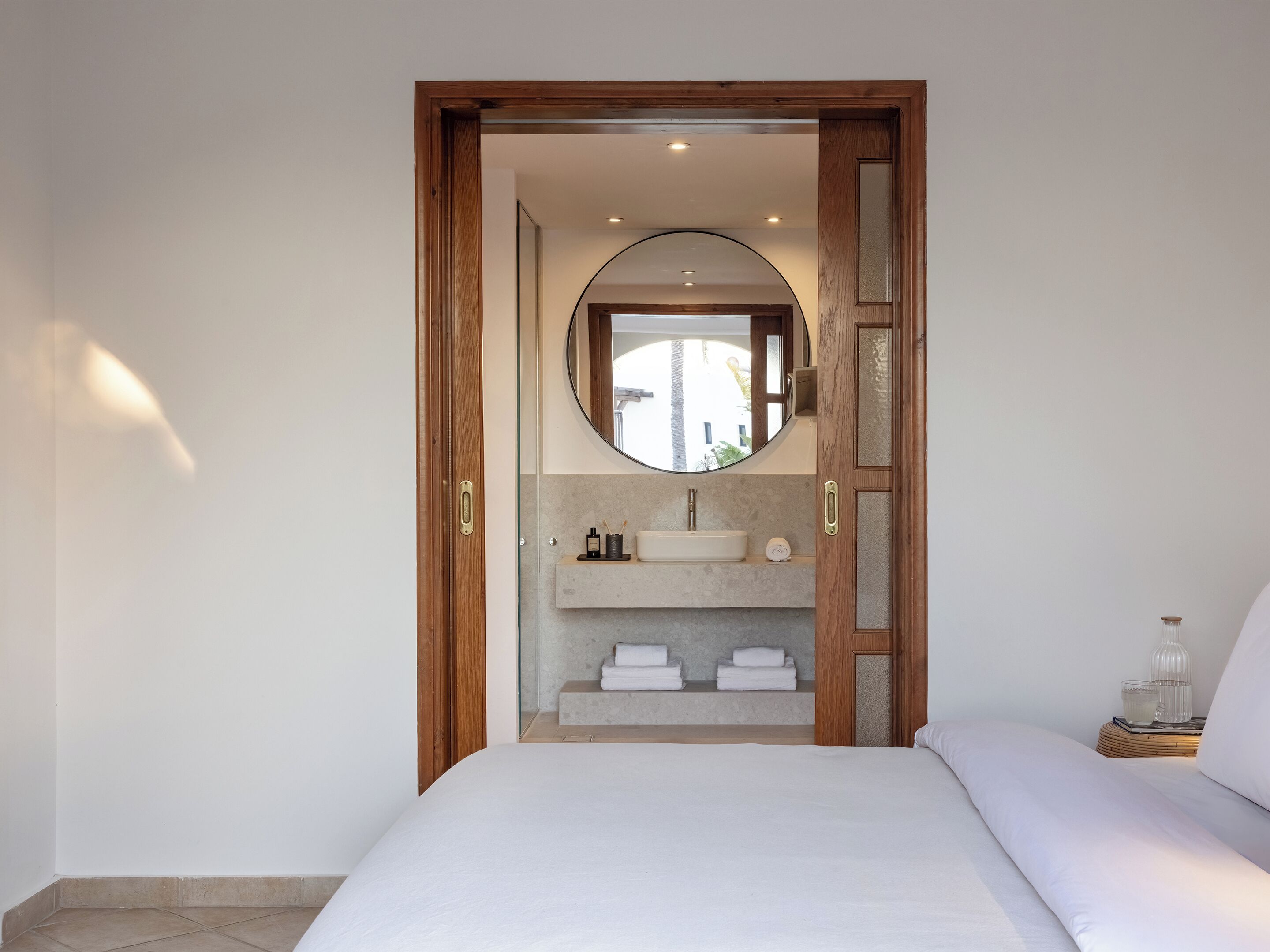 A bedroom with a bed, a wooden door leading to a bathroom with a sink and mirror.