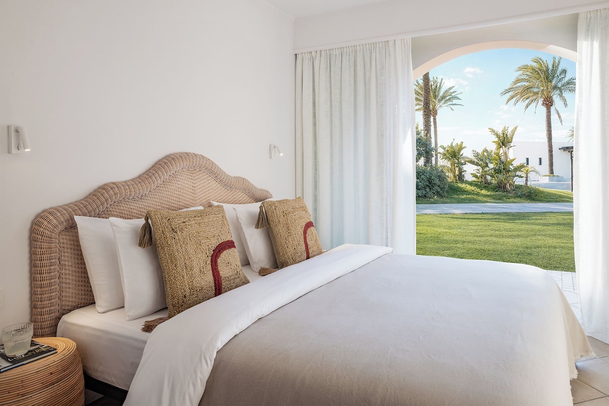 A bedroom with a large bed, a nightstand, and a view of a garden and palm trees.