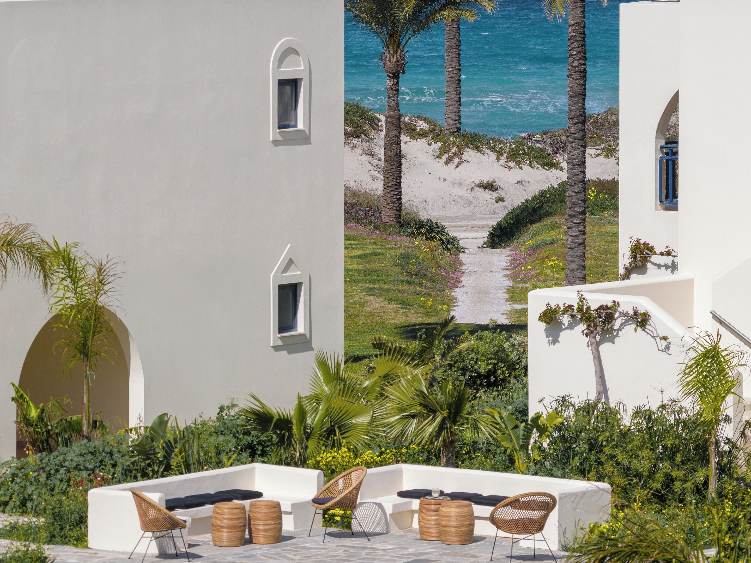 A white building with a view of the sea, palm trees, and a pathway leading to the beach.