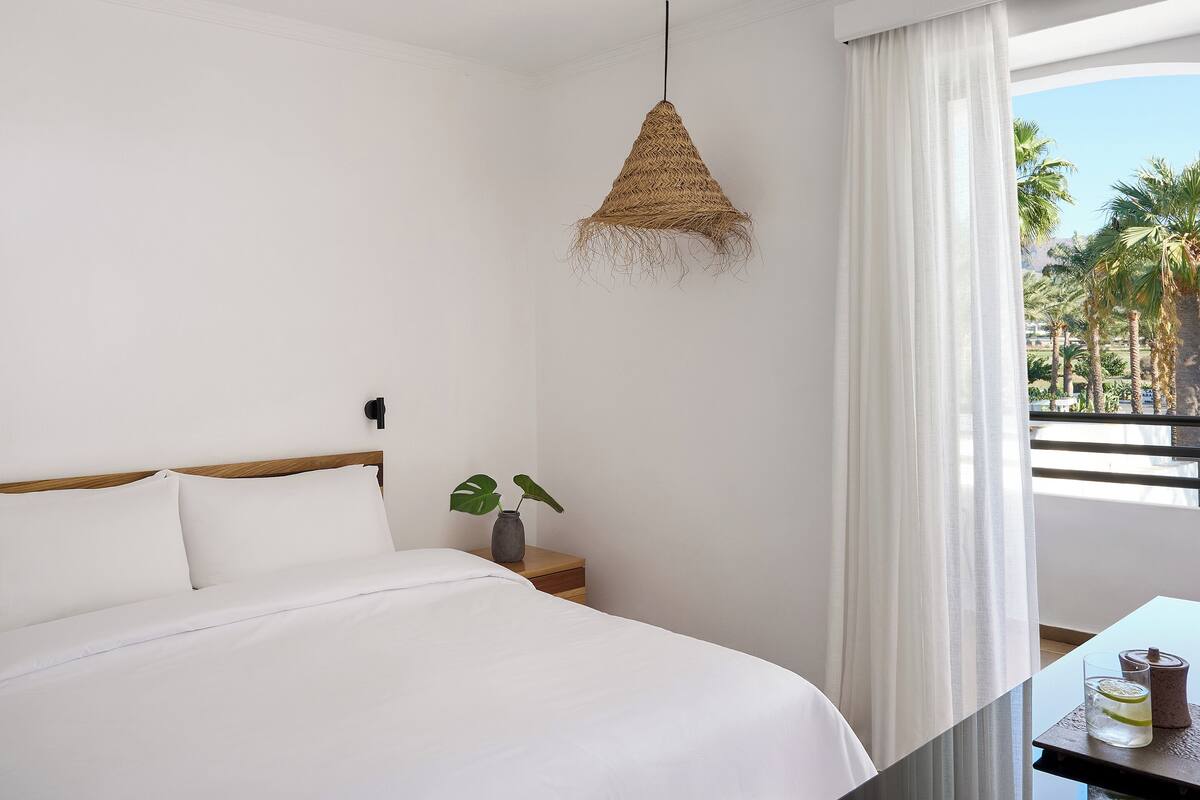 A neatly made bed with white linens, a bedside table with a plant, and a hanging woven light fixture.