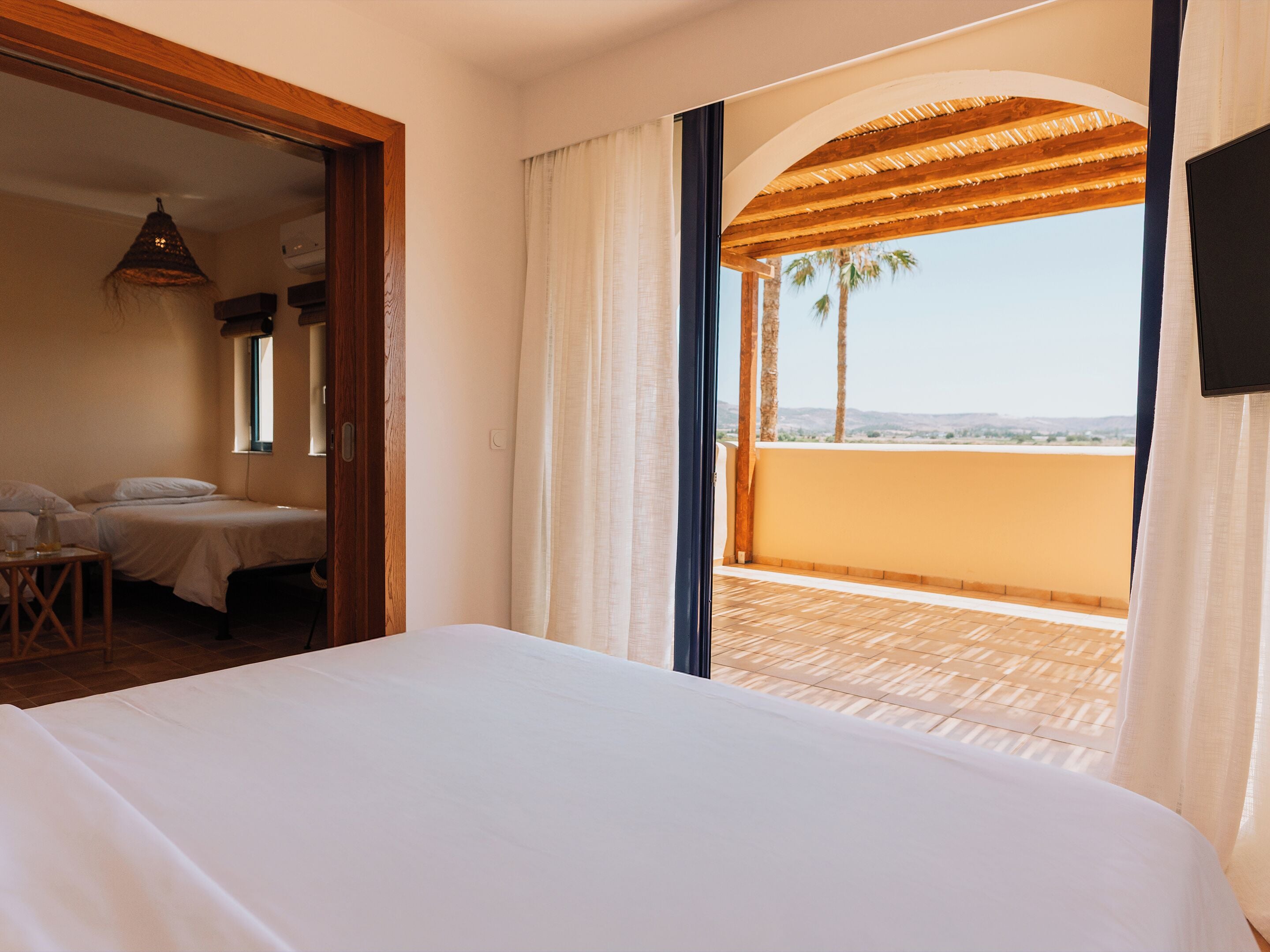A hotel room with a bed, a small table, and a chair. There is a balcony with a view of palm trees and a clear sky.