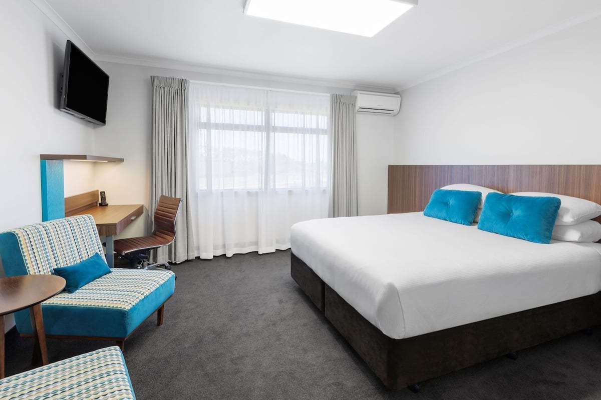 A hotel room with a bed, a desk with a chair, a TV mounted on the wall, and an air conditioning unit.