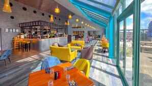 A modern restaurant with a bar area, yellow and blue seating, and a large glass ceiling.