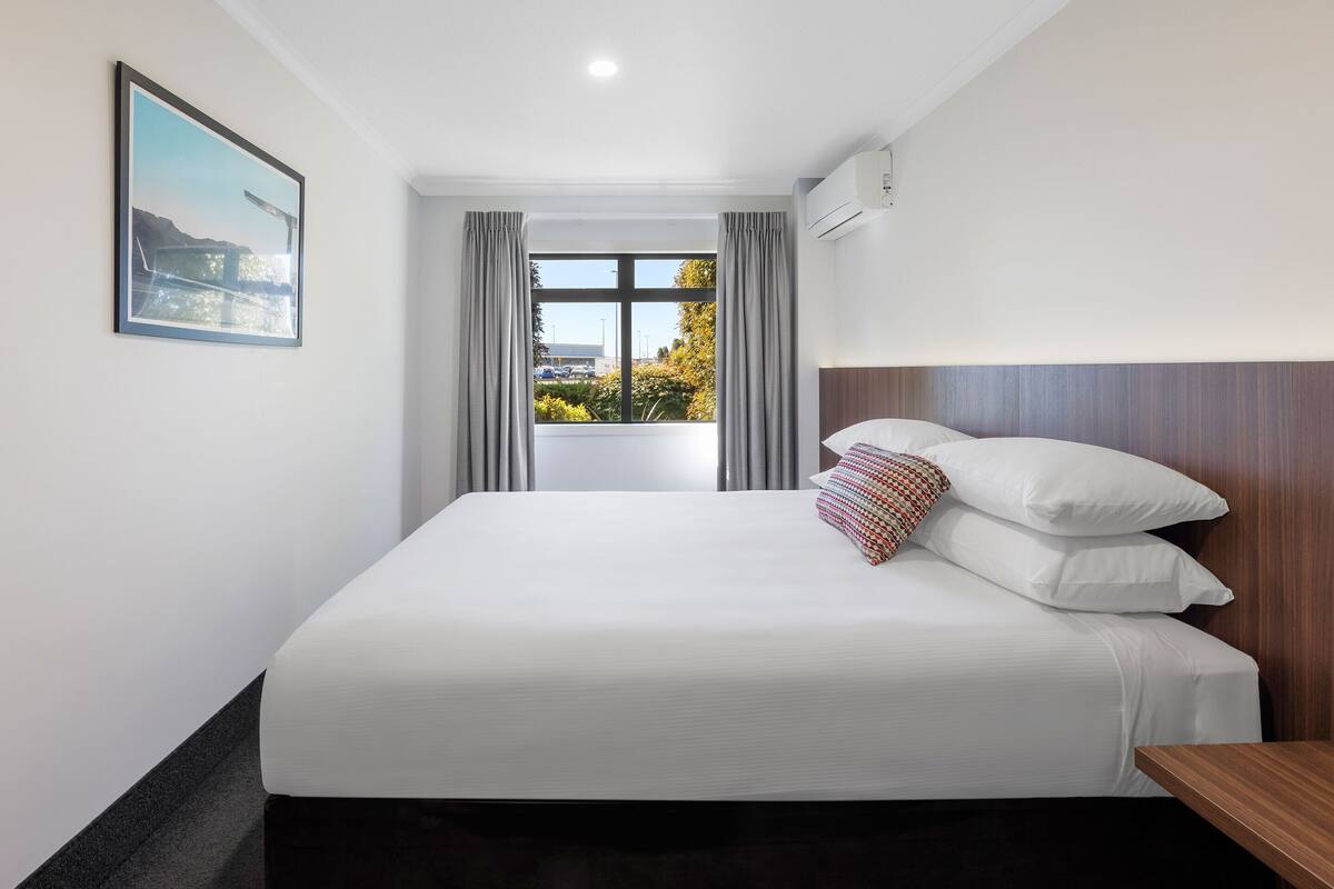 A hotel room with a large bed, a wooden headboard, a window with curtains, and a framed picture on the wall.