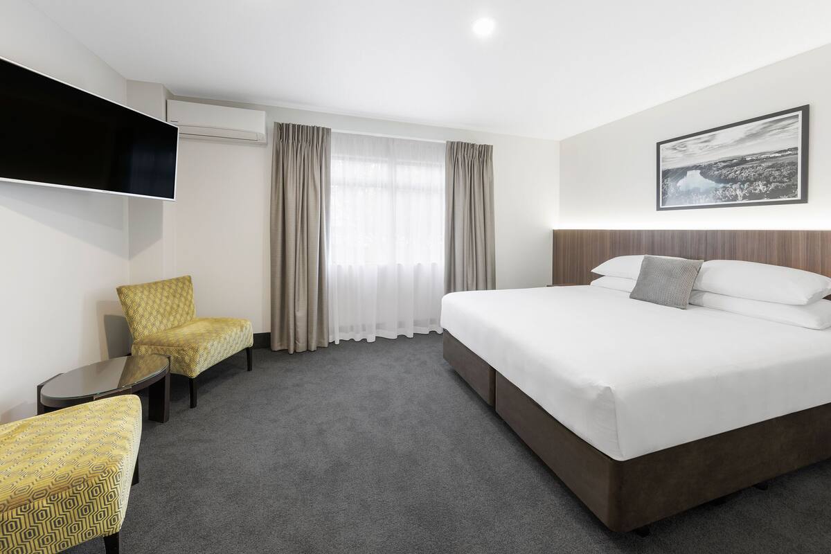 A hotel room with a large bed, a flat-screen TV, a chair, a small table, and a window with curtains.