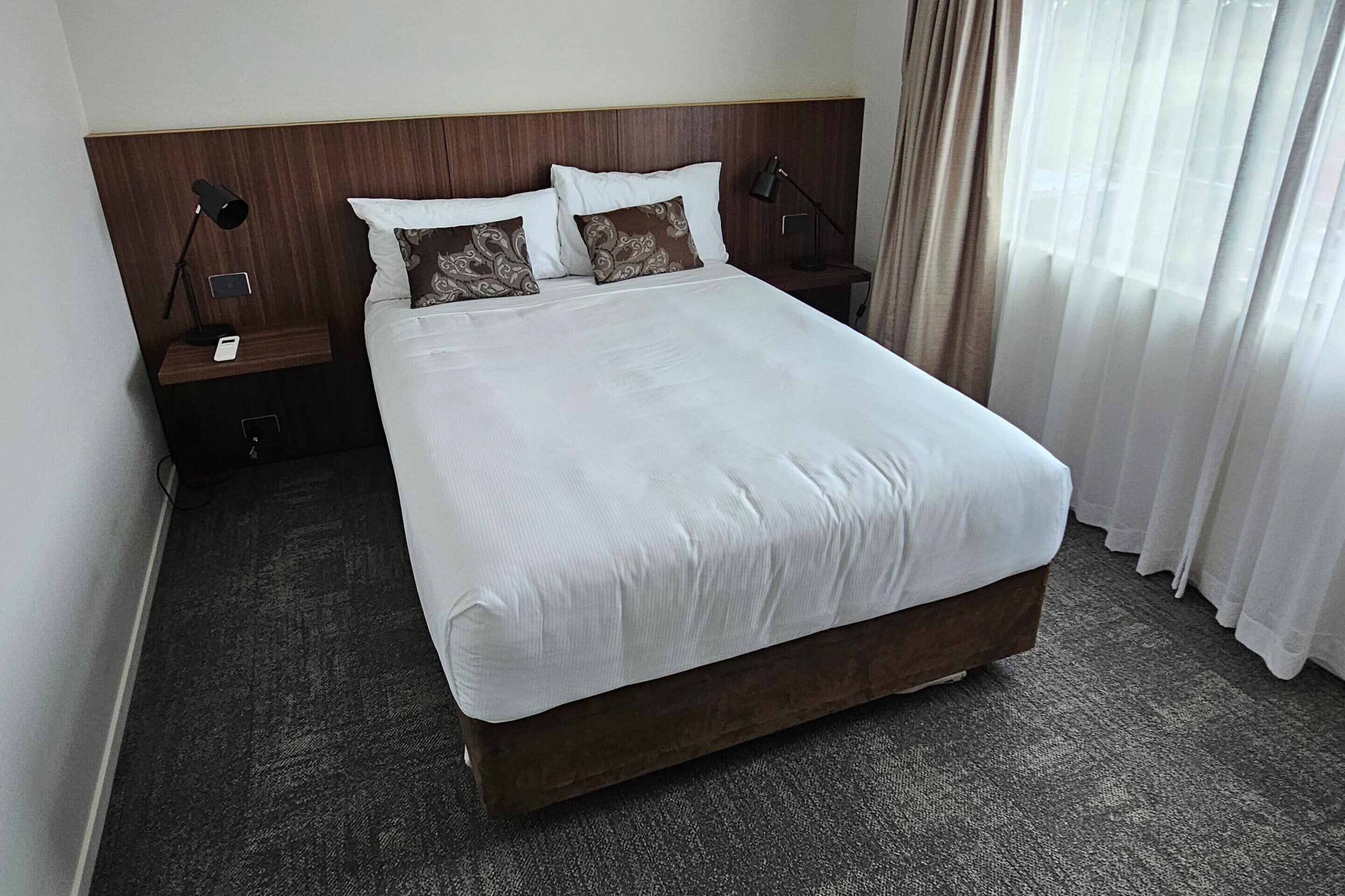 Habitación de hotel con una cama grande, dos mesitas de noche con lámparas, un cabecero de madera y vistas a una ventana con cortinas.