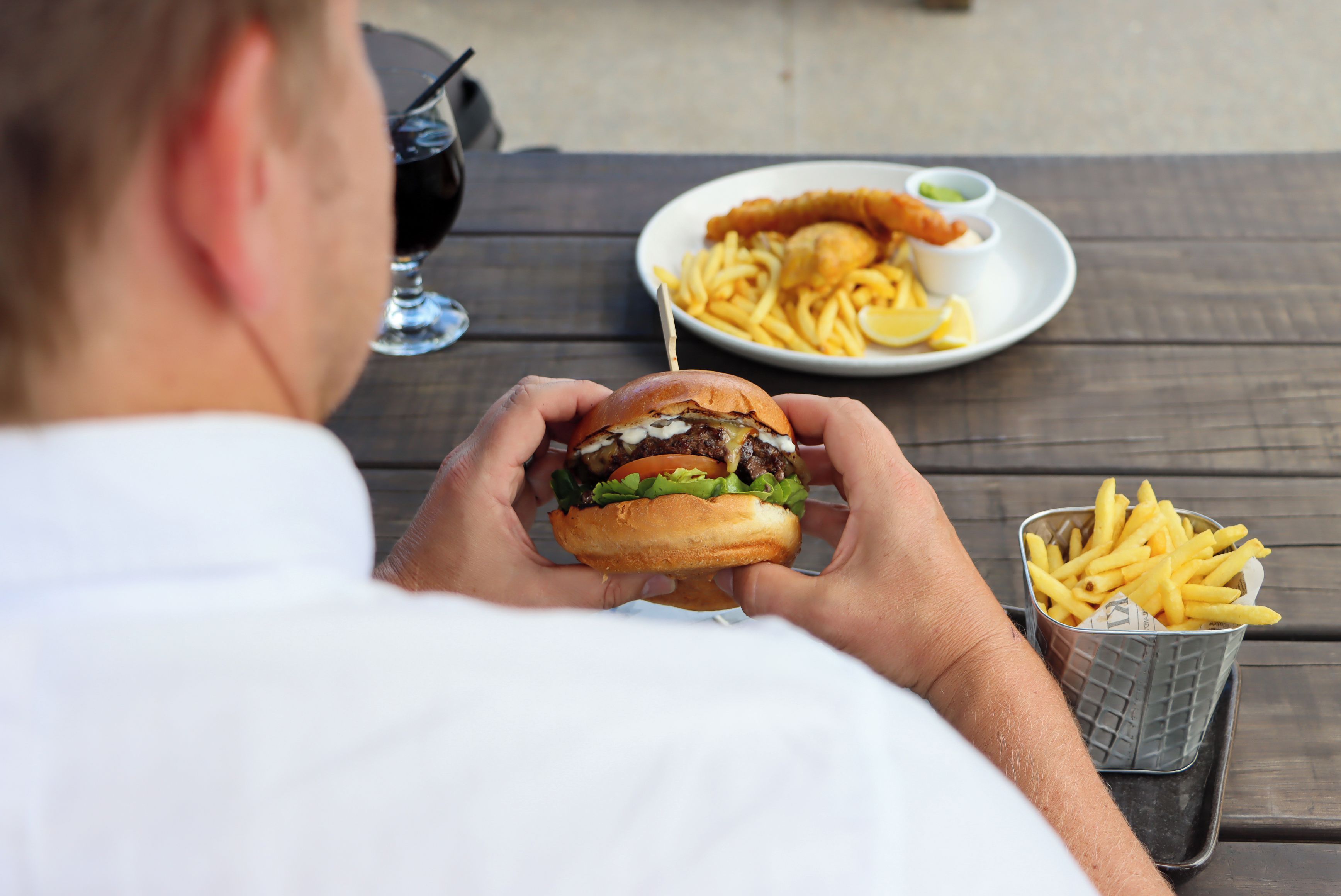 A person holding a burger with fries and a drink on a wooden table.