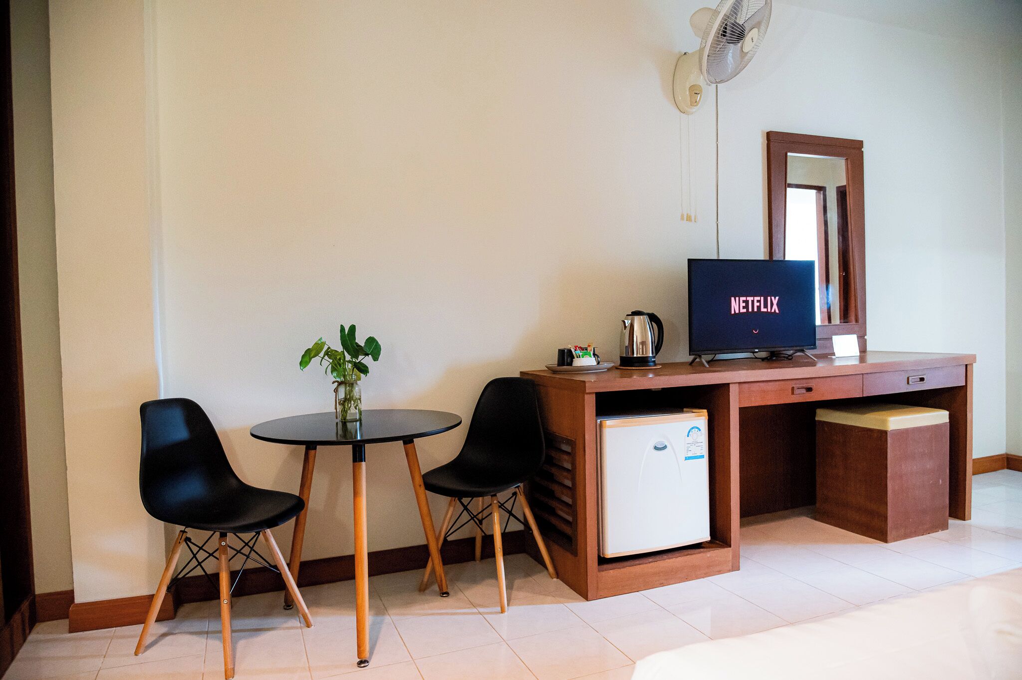A compact room with a wooden desk, a small refrigerator, a television, two black chairs, and a small table with a plant.