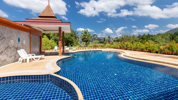 Ein Schwimmbecken mit blau gefliesten Rändern, Liegestühlen und einem Gebäude mit spitzer Dachform.