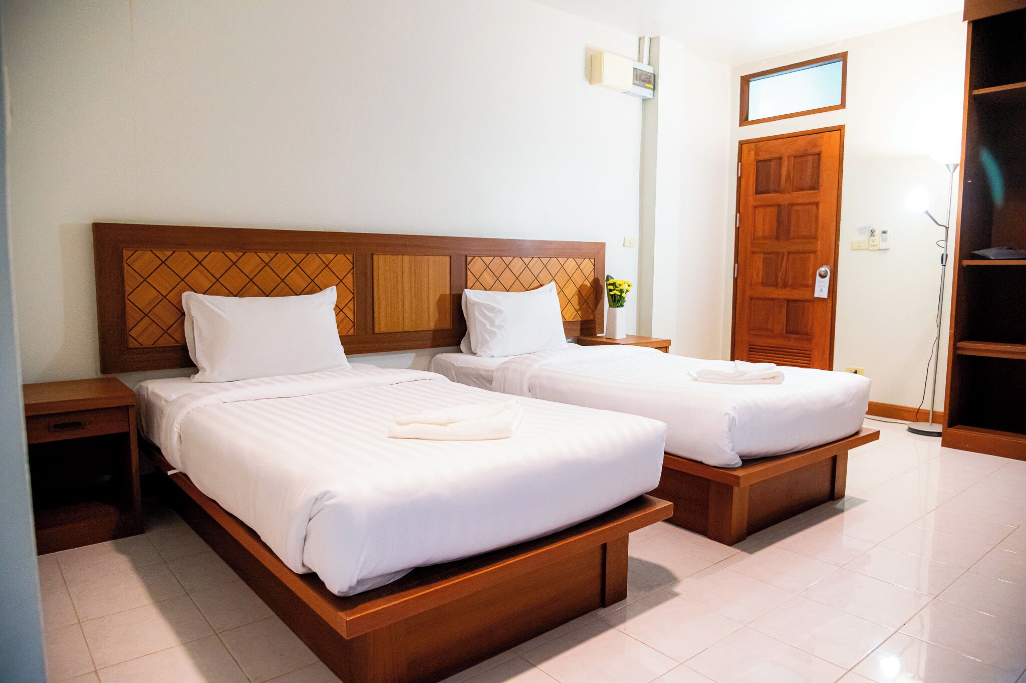 A hotel room with two single beds, wooden headboards, a bedside table with a lamp, and a door with a mirror above it.