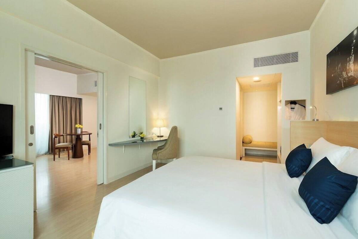 A hotel room with a bed, bedside table, lamp, chair, desk, television, and a door leading to another room.