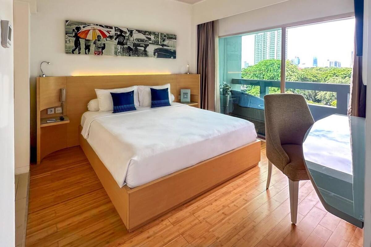 A modern hotel room with a large bed, a chair, a desk, and a balcony with a view of the city.
