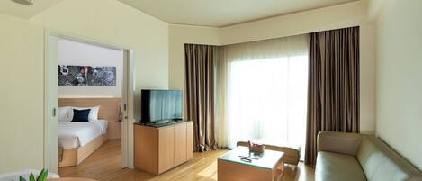 A modern hotel room with a bed, a TV on a wooden cabinet, a sofa, a coffee table, and a dining table with flowers.