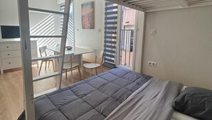 A room with a bed, a desk, and a chair. There is a window with blinds, a painting on the wall, and a staircase visible through the window. - Rent Cannes Meublés Gambetta (Cannes)