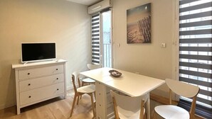 A modern, well-lit room with a dining table, chairs, a TV on a cabinet, and large windows with blinds. - Rent Cannes Meublés Gambetta (Cannes)