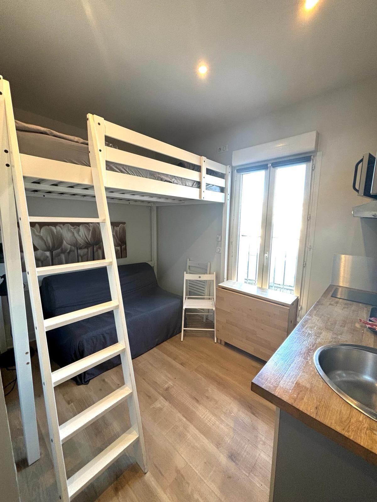 Habitació compacta amb un llit superposat, una cuineta petita, una finestra i una cadira.