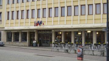 A hotel with a yellow facade, multiple windows, and outdoor seating with potted plants. Flags are displayed above the entrance.