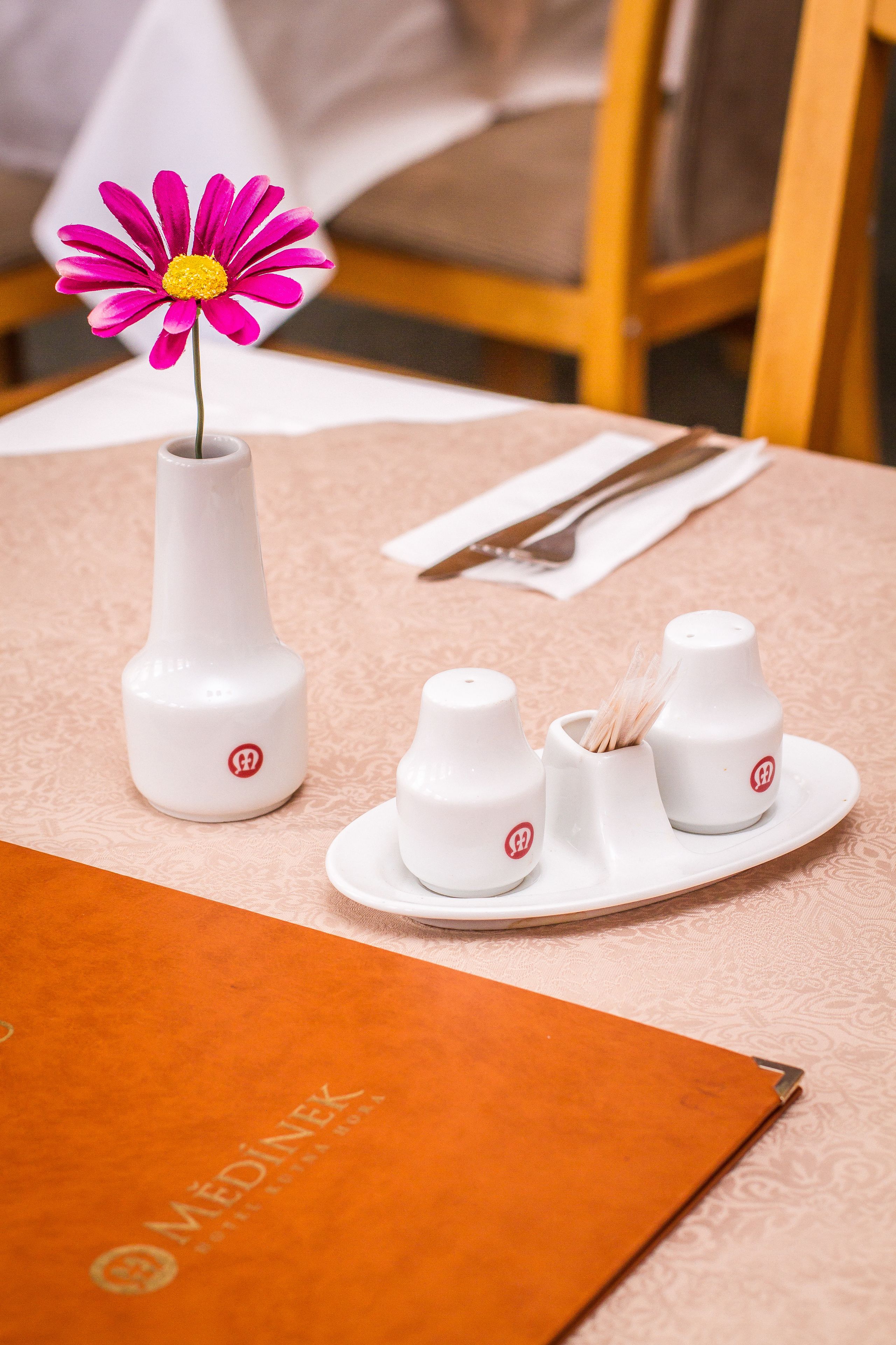 A table with a white vase containing a pink flower, two salt shakers, and a white napkin with cutlery.