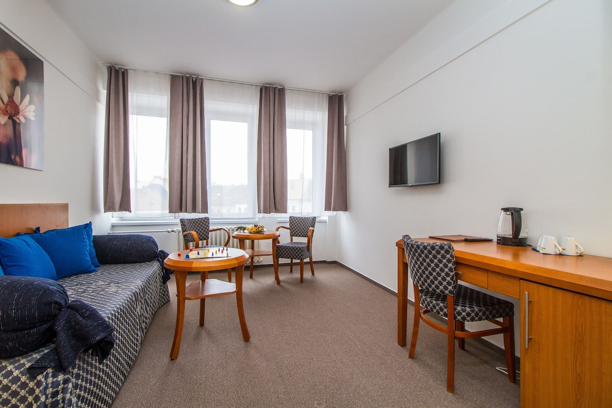 Habitación de hotel con sofá, dos sillones, una mesita, escritorio, televisor y ventana con cortinas.
