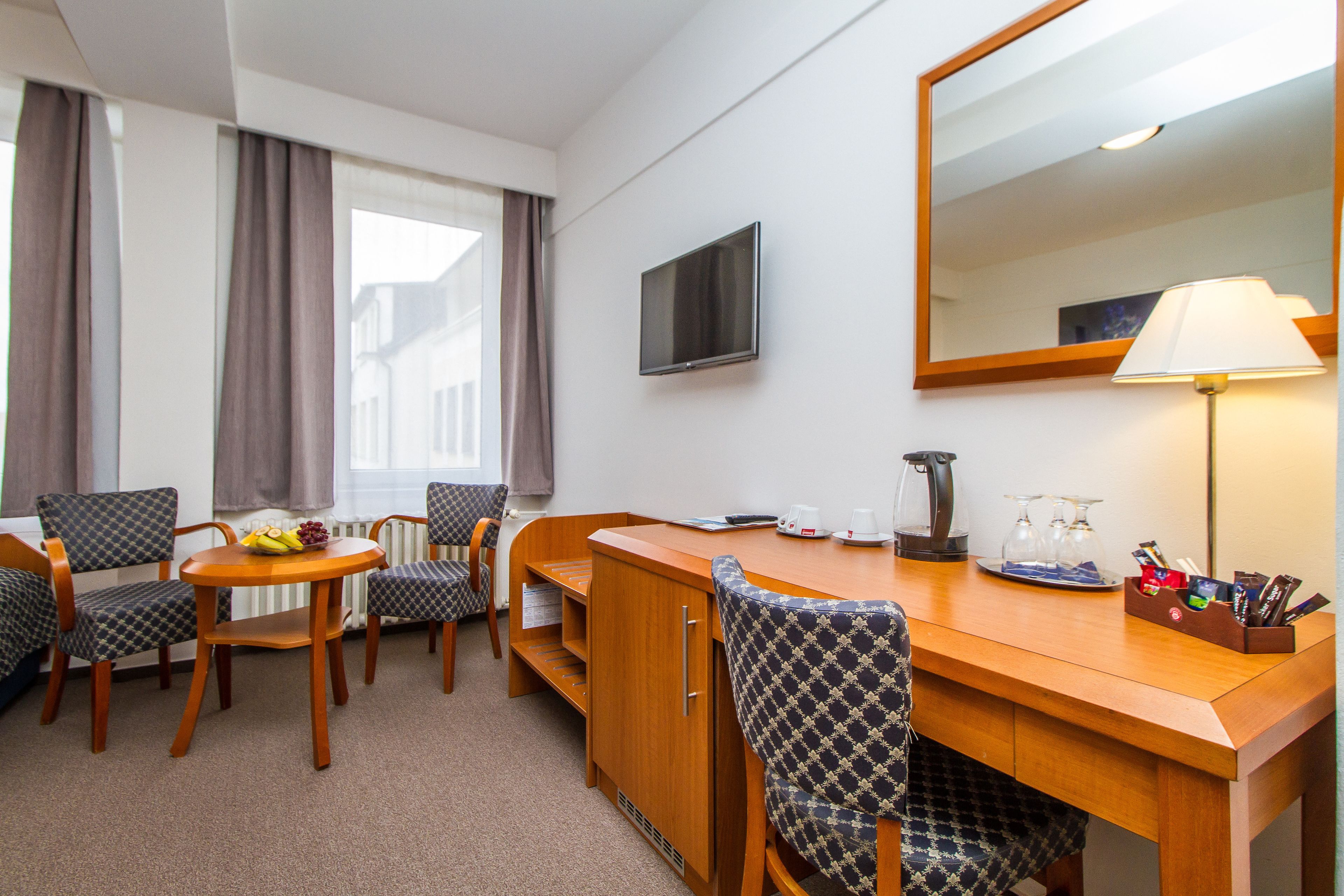A hotel room with a wooden desk, two chairs, a television, a lamp, and a window with curtains.