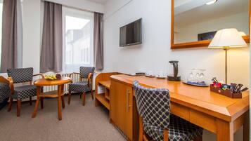 A hotel room with a wooden desk, two chairs, a television, a lamp, and a window with curtains.