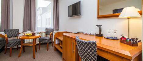 Habitación de hotel con escritorio de madera, dos sillas, televisor, lámpara y ventana con cortinas.