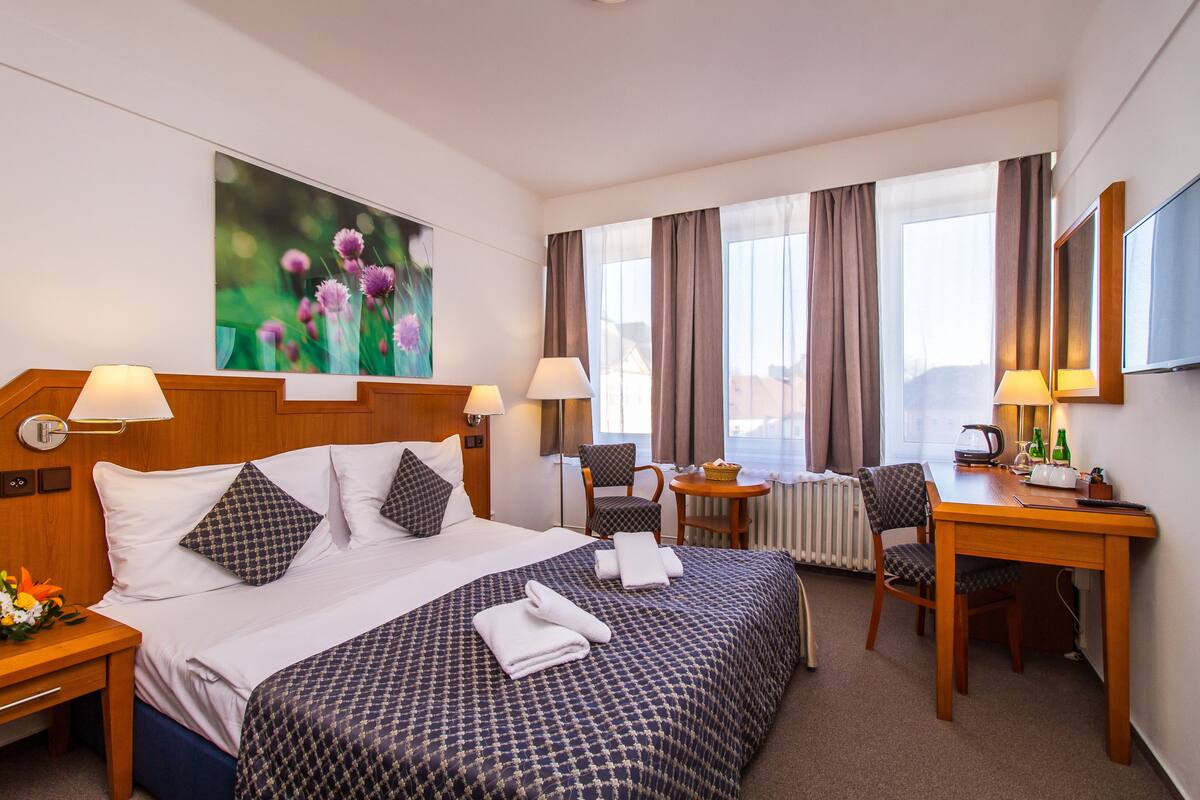 Una habitación de hotel con una cama grande, dos mesitas de noche con lámparas, un escritorio con una silla, un radiador y un ventanal con cortinas.
