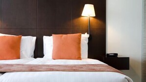 Una cama bien hecha con almohadas blancas y anaranjadas, un cabecero oscuro y una mesita de noche con una lámpara.