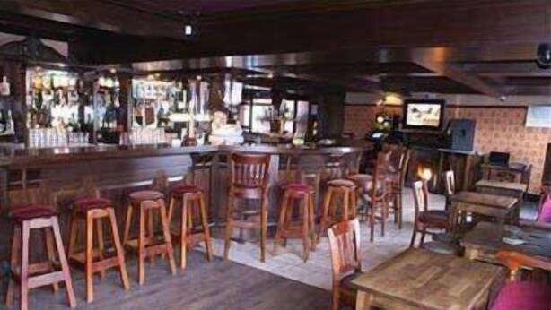 Una zona de bar con taburetes y mesas, una barra con televisor y una pared de ladrillo al fondo.