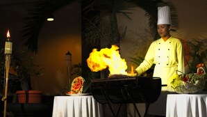 A chef performing a flambé with a flaming dish on a grill, surrounded by tables with floral centerpieces and a palm tree backdrop. - Singgasana Hotel Makassar (Makassar)