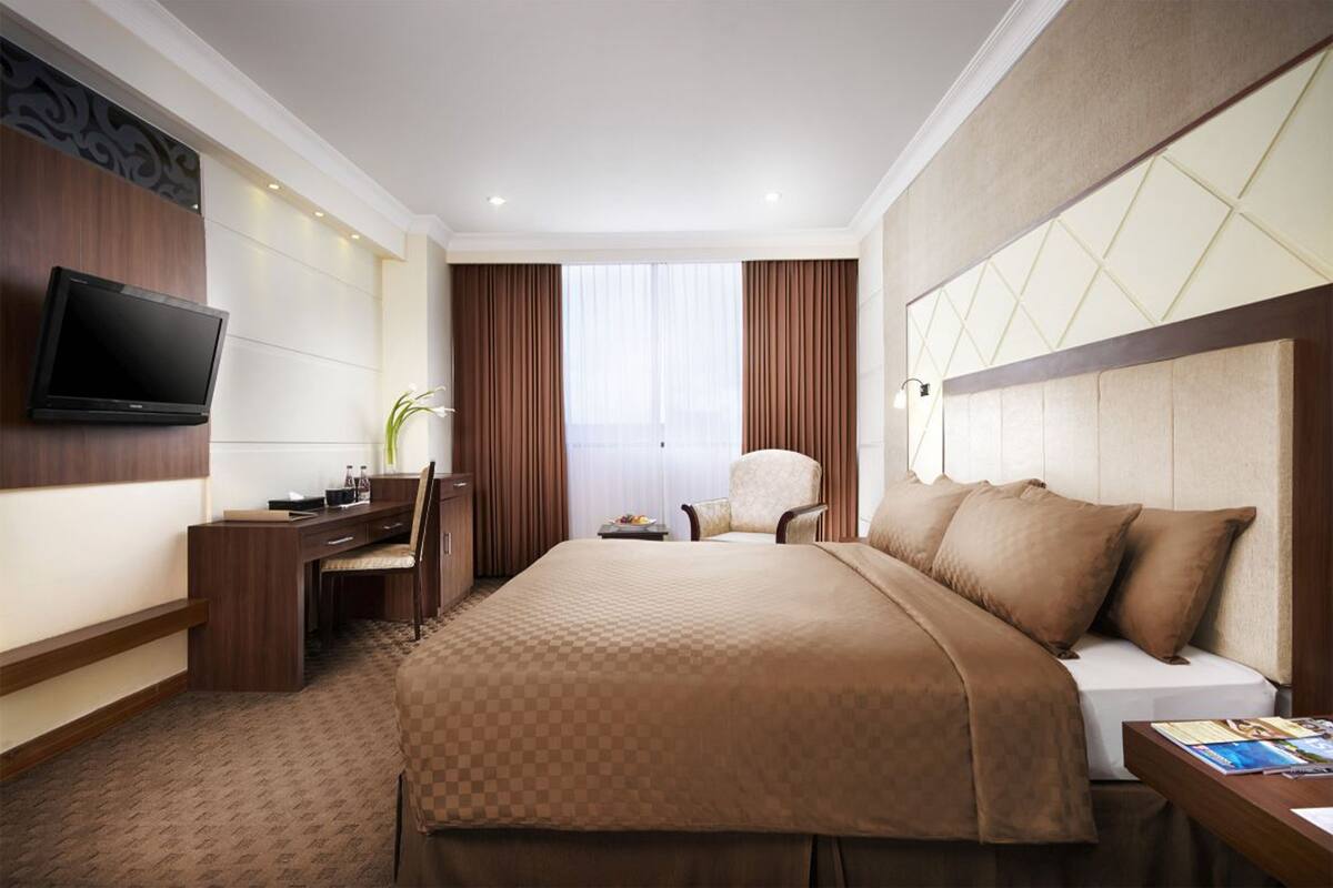 A hotel room with a large bed, a desk with a chair, a flat-screen TV mounted on the wall, and a window with curtains.