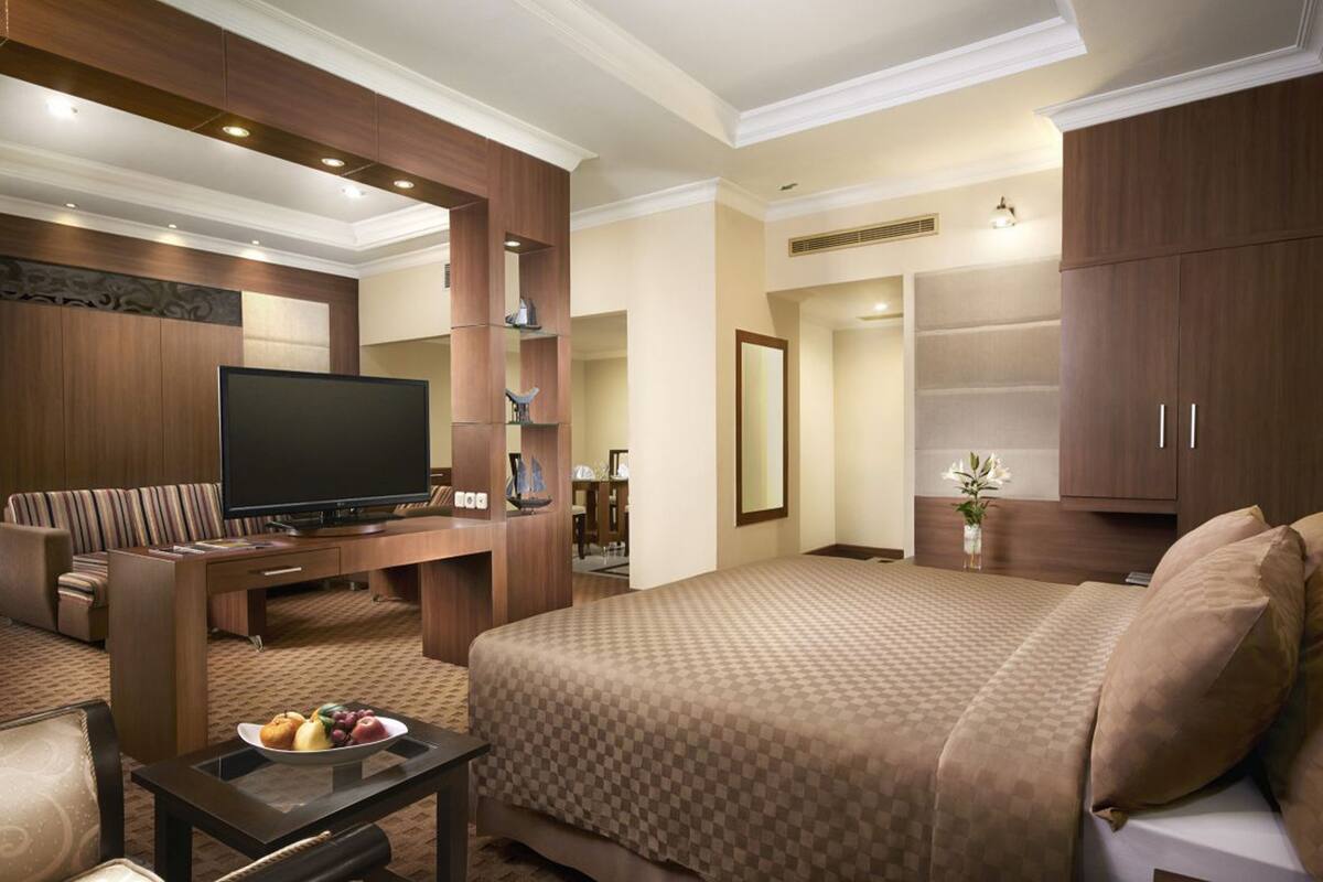 A hotel room with a large bed, a flat-screen TV, a seating area with a sofa and armchairs, and a dining table with fruit.