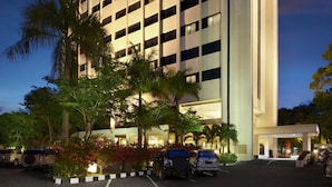 A multi-story hotel with a prominent sign at the top, a parking area with several cars, and a well-lit entrance. - Singgasana Hotel Makassar (Makassar)