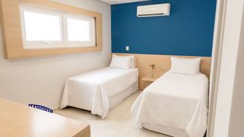 A hotel room with two single beds, a wooden desk, a small bedside table with a vase, and a window with blinds.