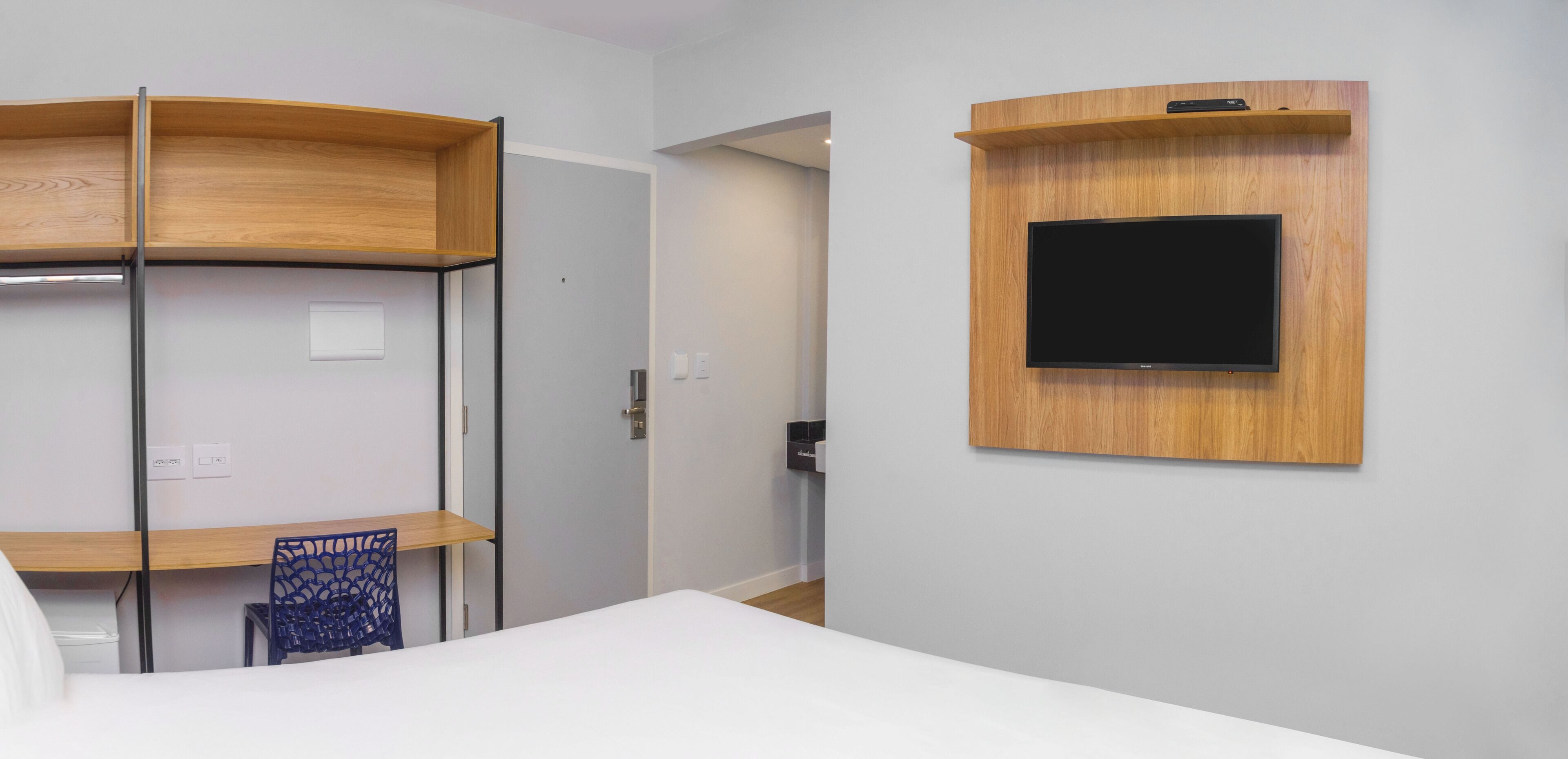 A hotel room with a bed, a desk with a chair, a TV mounted on the wall, and a door leading to another room.