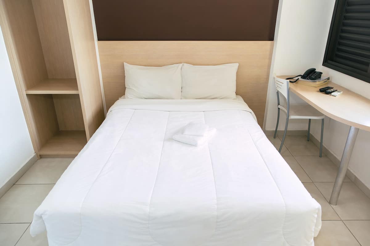 A neatly made bed with white bedding, a wooden headboard, a desk with a telephone, and a chair.