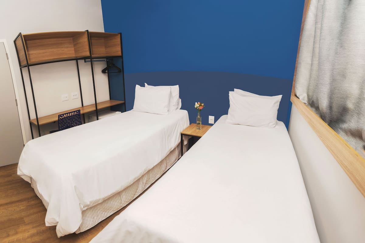 A room with two single beds, a wooden shelf unit, a small table with a vase, and a blue accent wall.