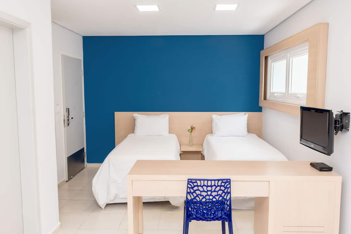 A hotel room with two beds, a desk, a chair, a TV, and a window.