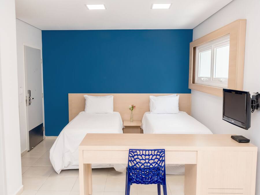 Ein Hotelzimmer mit zwei Betten, einem Schreibtisch, einem Stuhl, einem Fernseher und einem Fenster.
