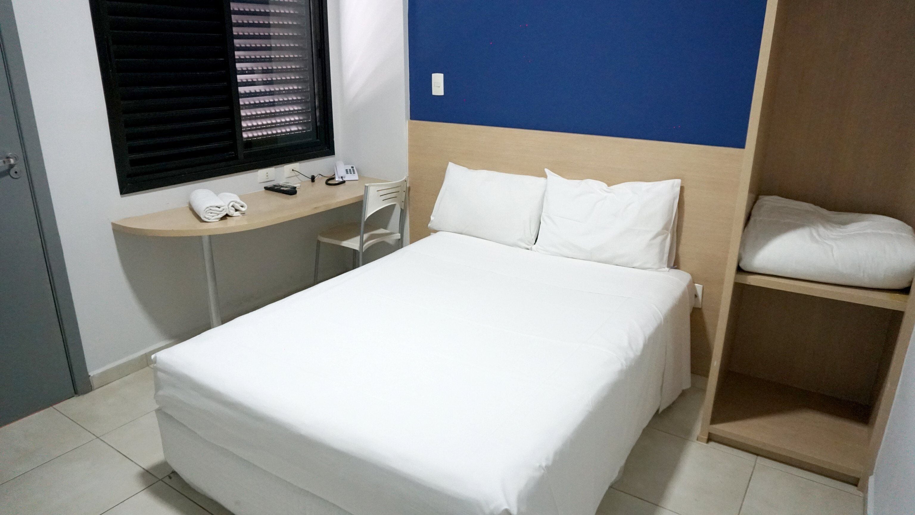 Una cama bien tendida con sábanas blancas, una mesita de noche con teléfono y control remoto, una ventana con persianas y una pared azul de acento.