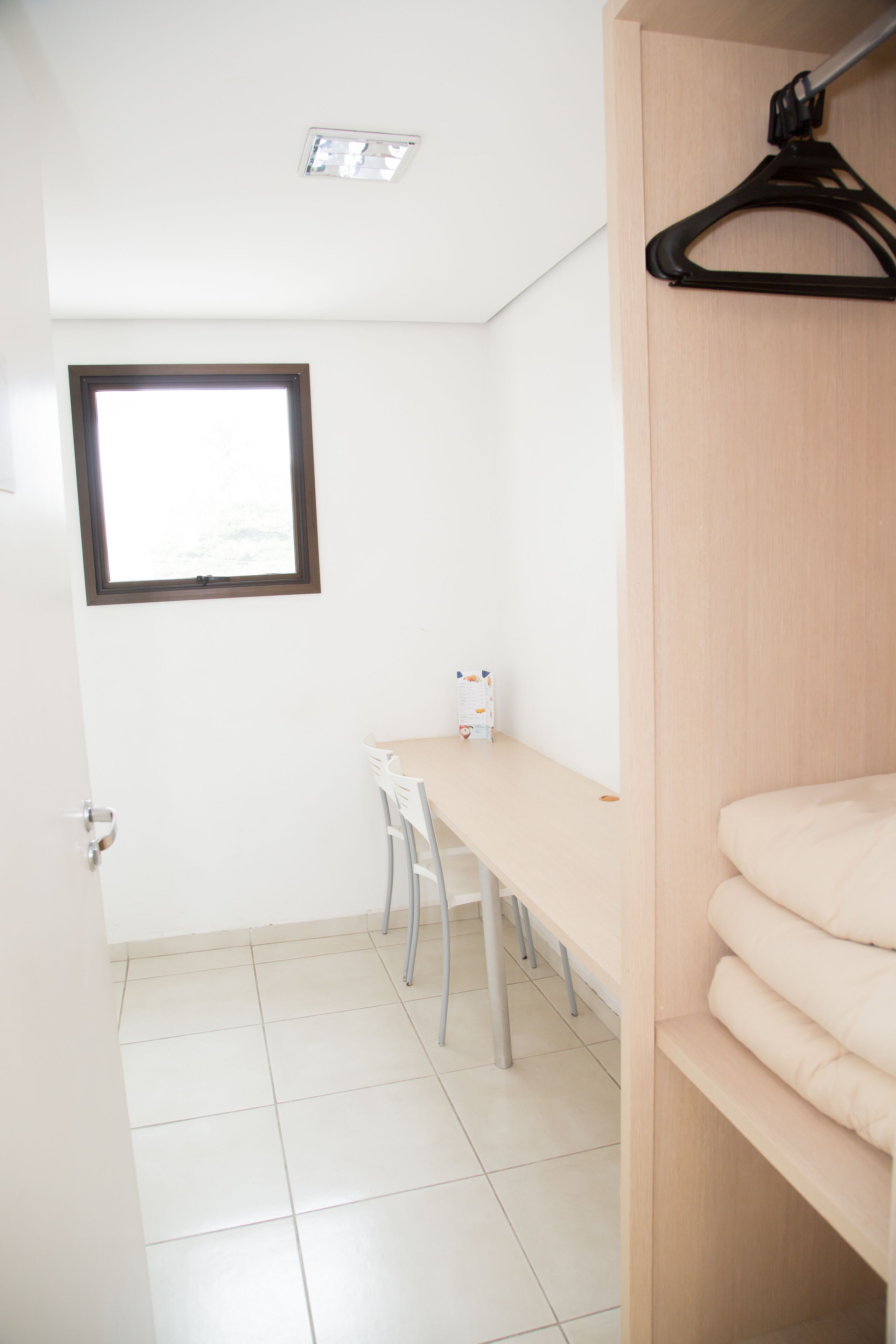 Una habitación pequeña y limpia con un escritorio de madera, una silla, una cama con cabecera de madera, un perchero y una ventana con persianas.