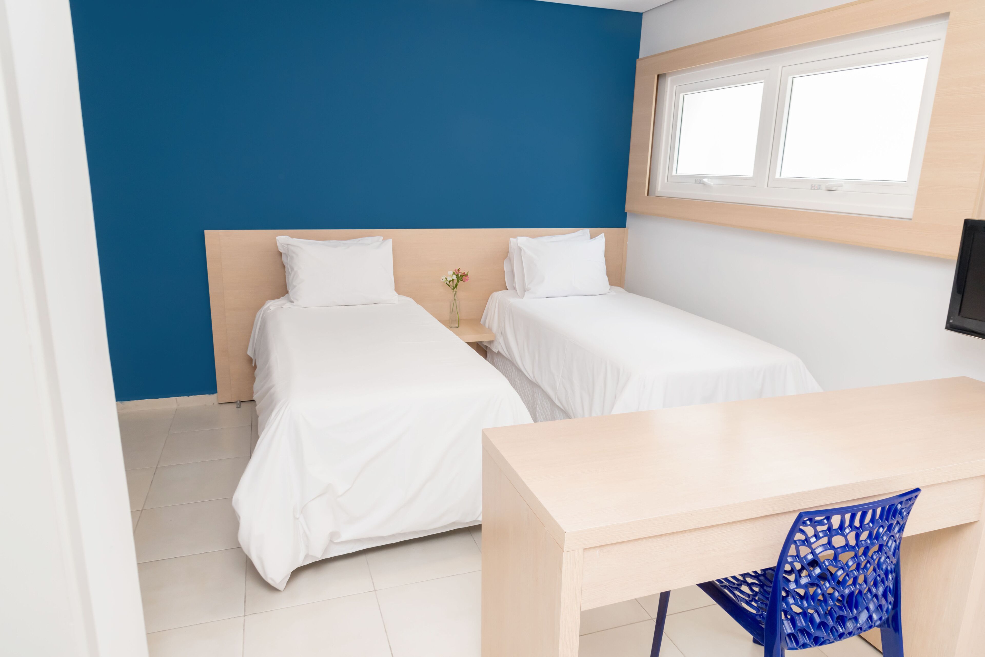 A hotel room with two single beds, a wooden desk, a blue chair, and a window with blinds.