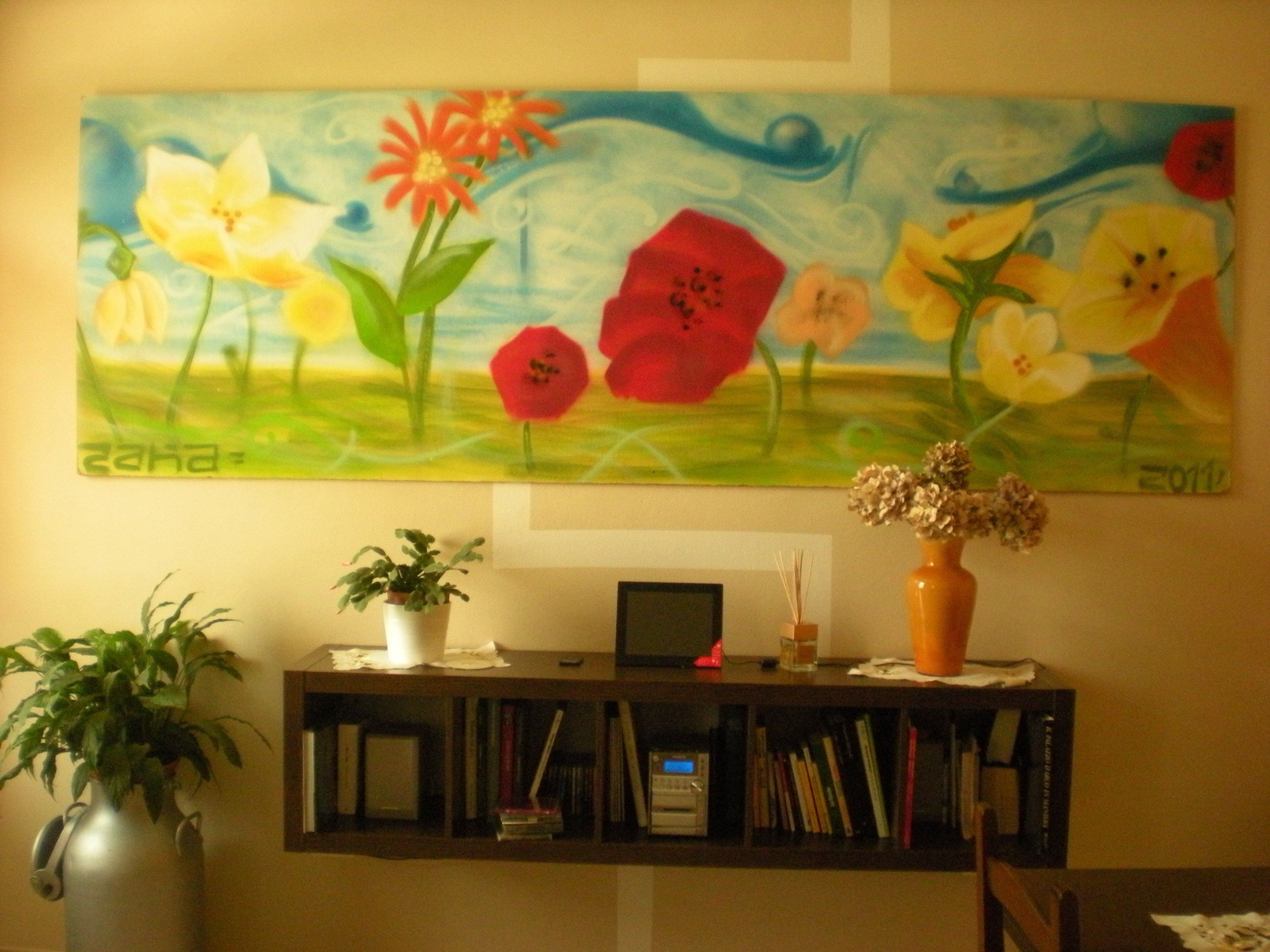 A painting of flowers on a wall, a bookshelf with books and a small plant, and a vase with dried flowers on a table.