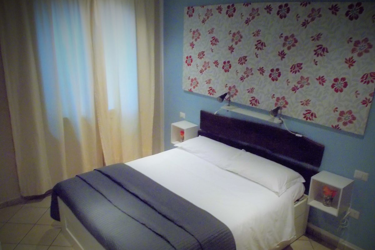Un lit bien fait, avec une tête de lit sombre, une décoration murale à motifs floraux au-dessus, et une petite table de chevet sur laquelle se trouve une lampe.