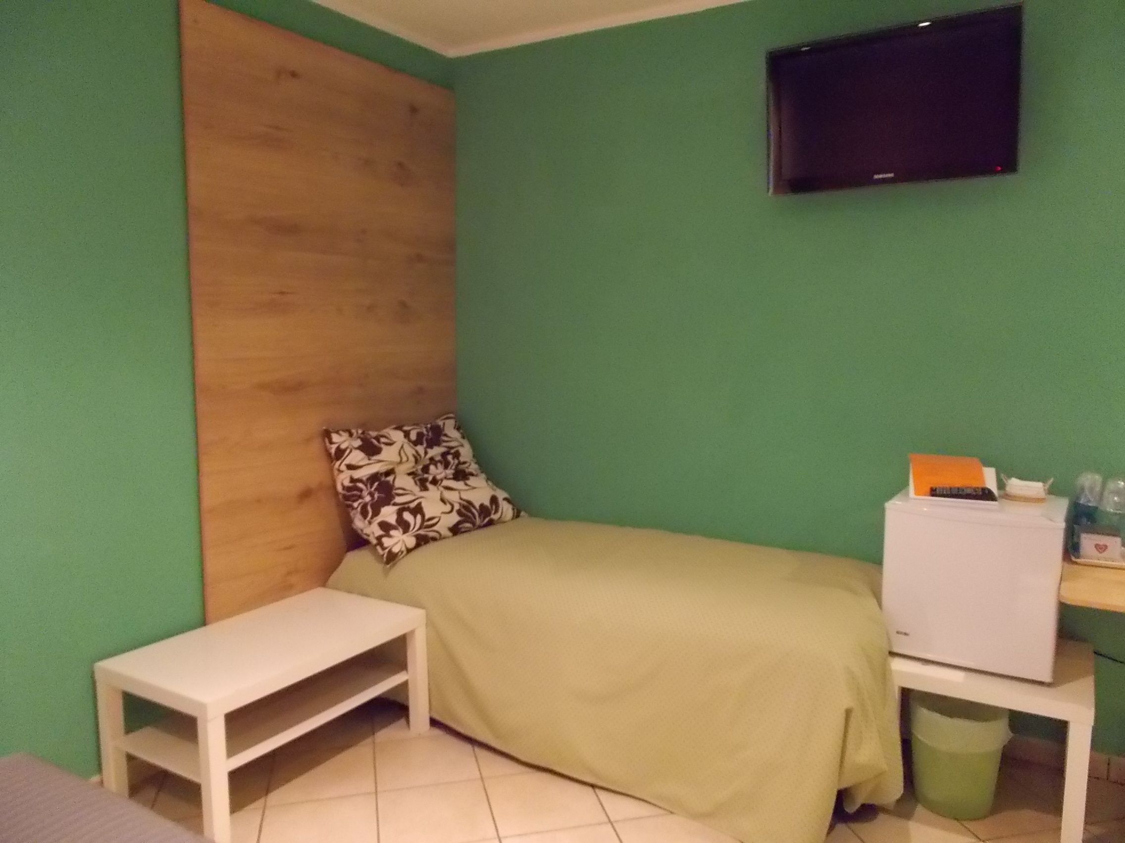 Ein kleines, ordentlich eingerichtetes Schlafzimmer mit Bett, Holz-Kopfteil, Wand-Fernseher, einem kleinen Tisch und einem Mülleimer.