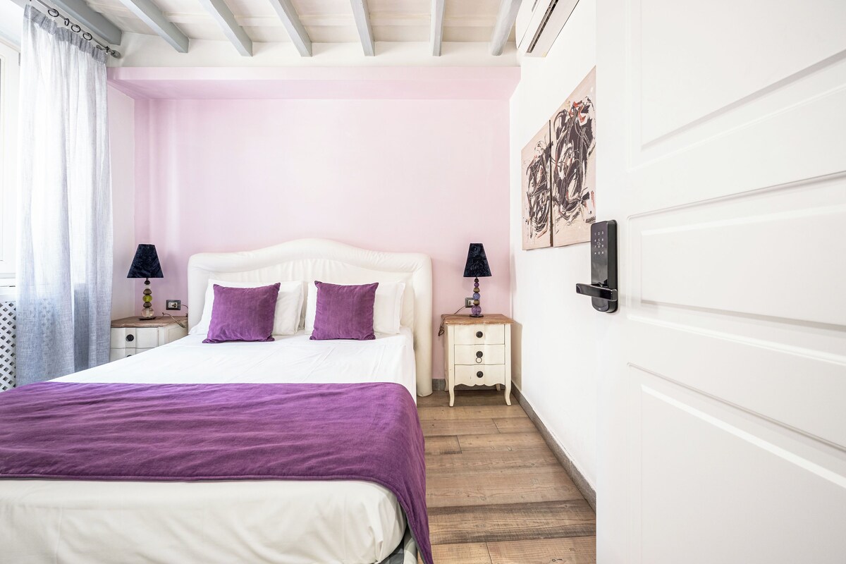 A bedroom with a bed, two bedside tables, a nightstand, a door, and a wall with artwork.
