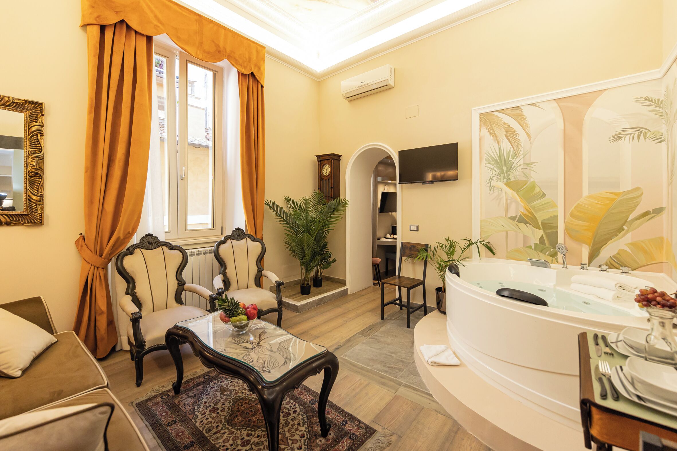 A living room with a sofa, armchairs, a coffee table, a dining area with a table set for two, a large window with curtains, a clock, and a wall-mounted TV.