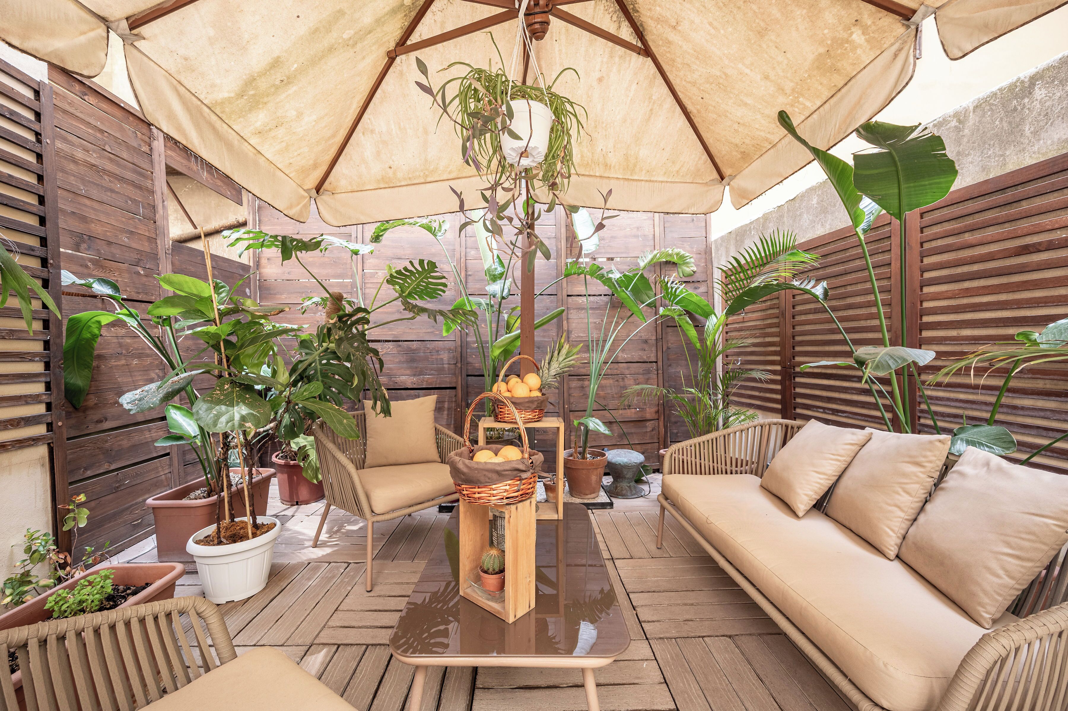 Un patio al aire libre con piso de madera, mobiliario de mimbre y diversas plantas en macetas.