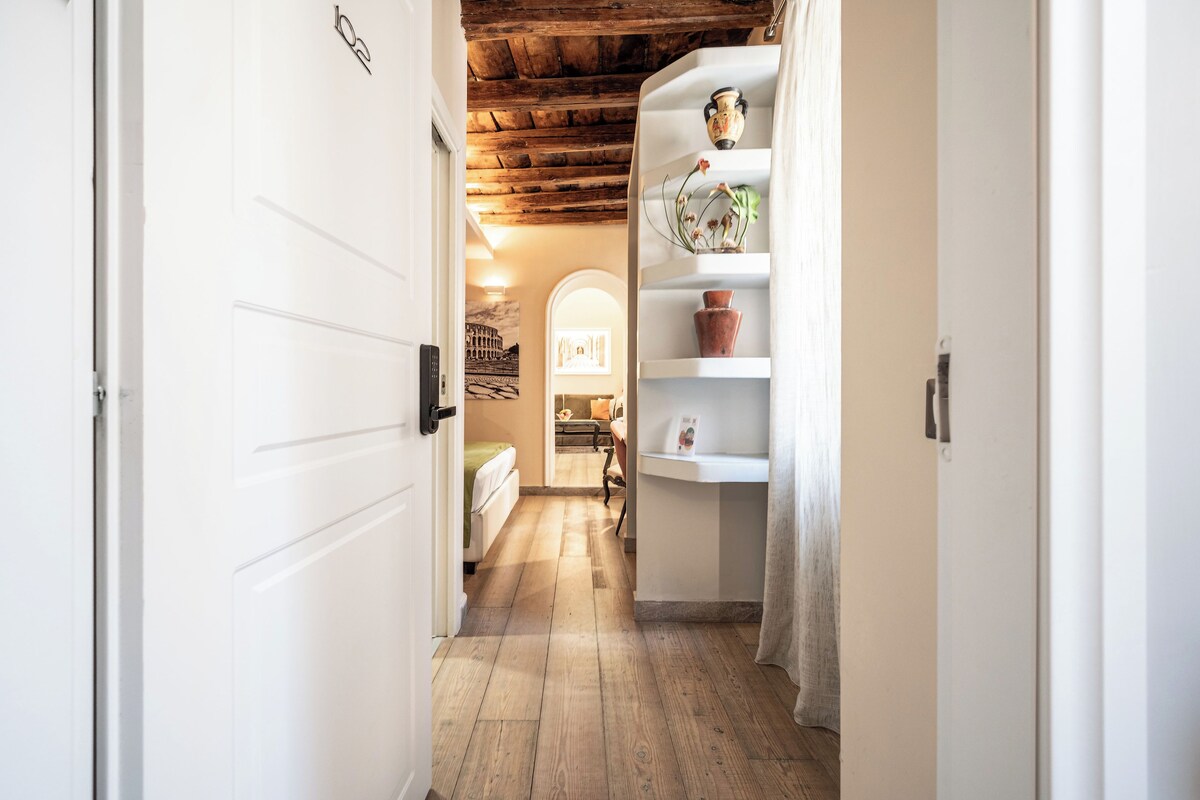 A hallway with a wooden floor, white walls, and a ceiling with exposed beams. There is a bed with a green blanket and a small table with a vase and a picture frame.